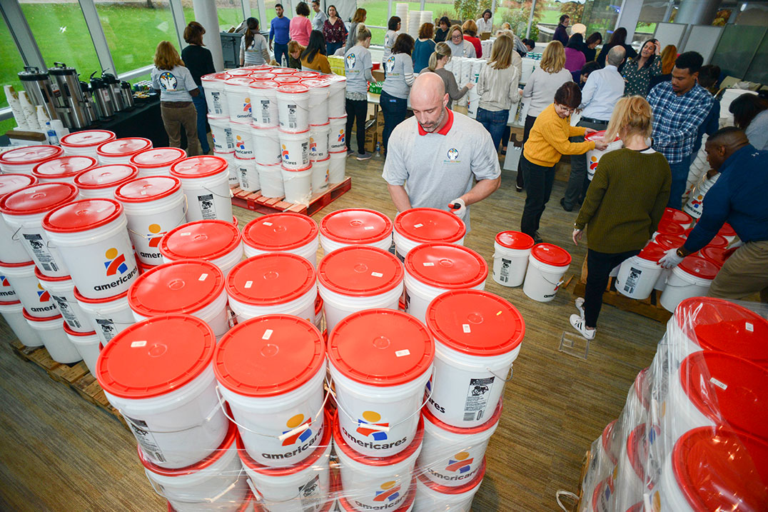 Stacked white buckets with orange lids and a group of volunteers in the background