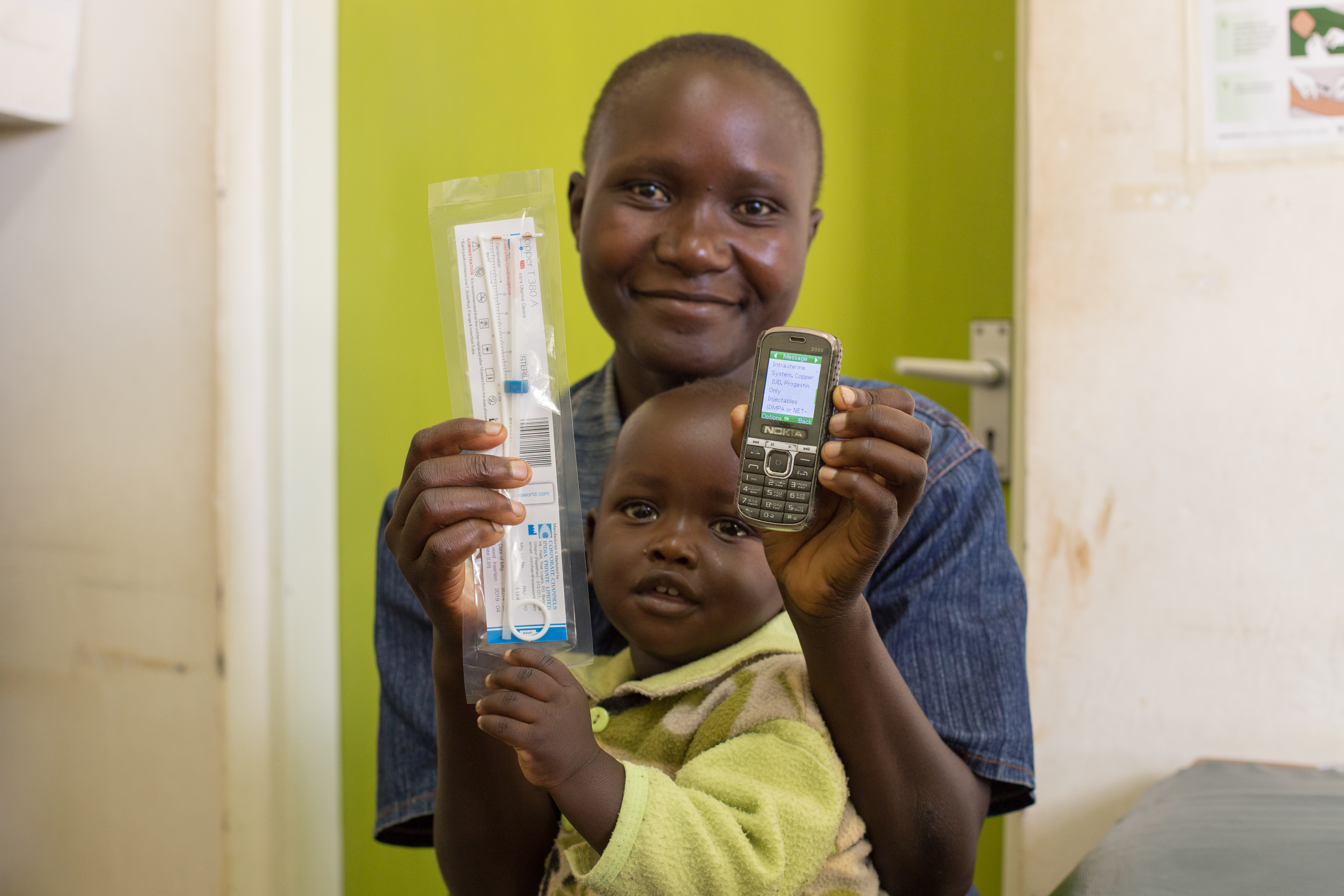 A smiling person holding a young child in front of a green door, displaying a packaged medical syringe in one hand and an old mobile phone in the other