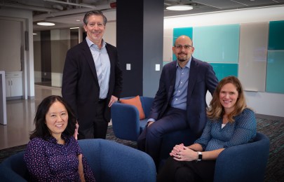 Four people in a modern office setting, with two seated on blue chairs and two standing behind them