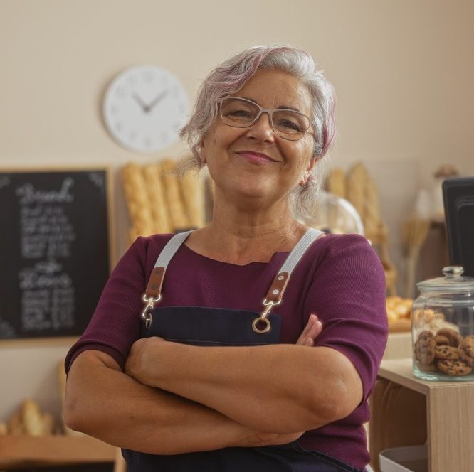 a person wearing an apron standing with arms across inside a bakery.