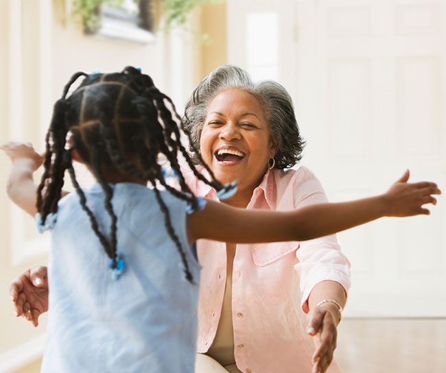 older person with pink shirt smiling outstretching her arms to hug a younger person with braids.
