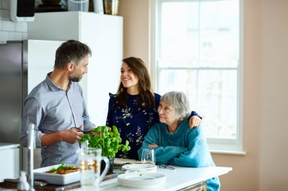 younger woman in the kitchen with her arm around an older woman and a young man nearby smiling